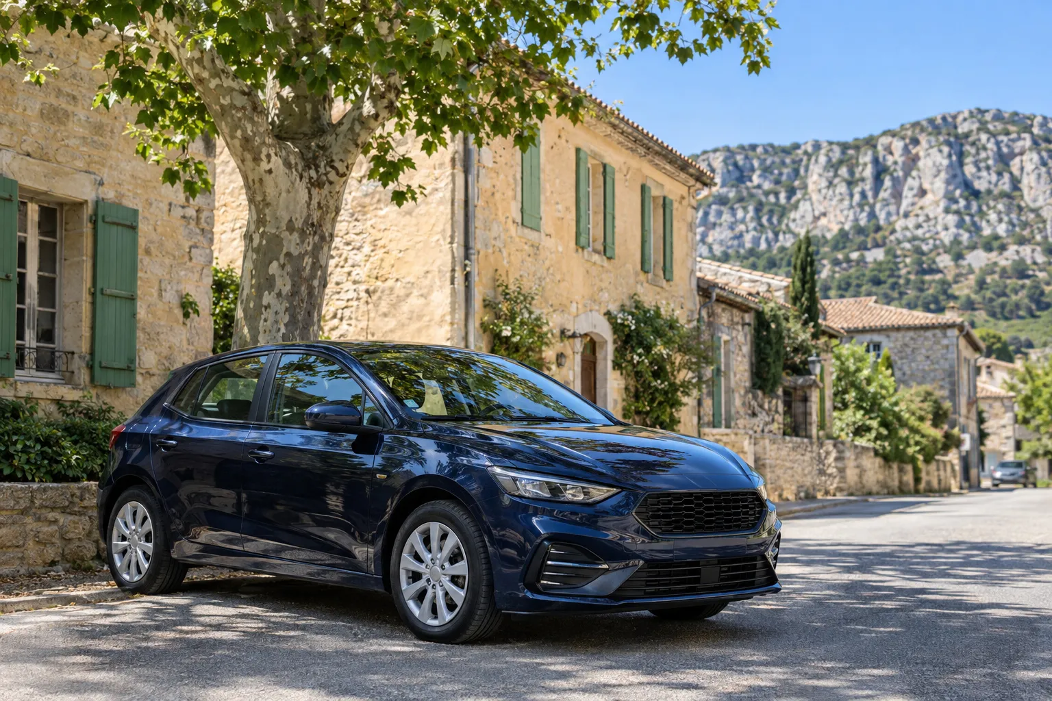 Une compacte bleu foncé garée à l’ombre d’un platane dans une rue de village provençal, maisons en pierre et volets verts, falaises calcaires au fond.